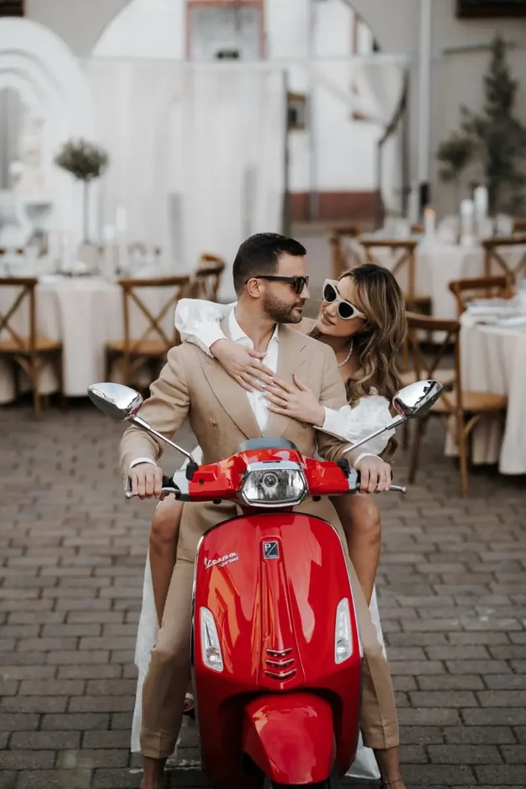 Paar in eleganter Kleidung auf rotem Vespa-Roller, lächelnd und entspannt, mit stilvoller Tischdeko im Hintergrund.