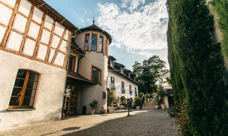 Historischer Fachwerkbau in sonniger Gasse mit grünen Zypressen und blauem Himmel.