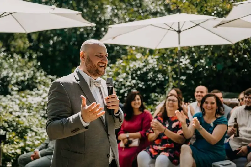 Mann hält Rede bei Hochzeit im Freien, umgeben von applaudierenden Gästen unter weißen Sonnenschirmen.