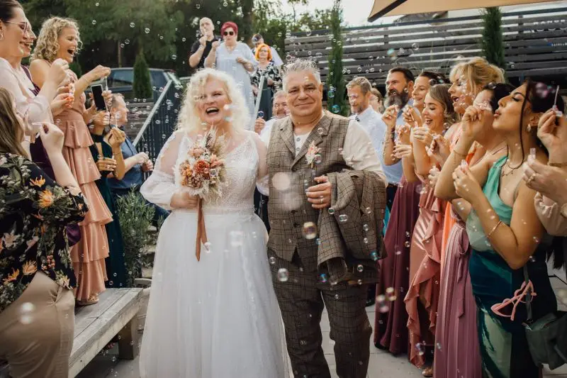 Brautpaar verlässt feierlich die Hochzeit unter einem Regen aus Seifenblasen, umgeben von applaudierenden Gästen im Freien.