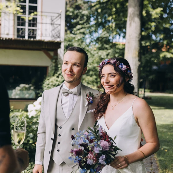 Brautpaar im Freien, lächelnd mit Blumenstrauß und Blumenkronen, bei sonniger Hochzeit im Garten.